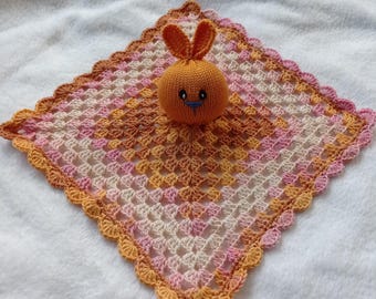 Puede incluir: Una manta de bebé de ganchillo en tonos rosa, crema y naranja con un juguete de conejo naranja de ganchillo en el centro.