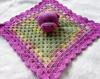 Puede incluir: Una manta de bebé de ganchillo de color morado con un juguete de cerdo de ganchillo morado en el centro. La manta tiene un borde festoneado y está hecha con una variedad de tonos de rosa, verde y amarillo.