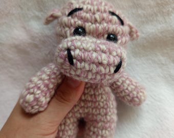 Puede incluir: Un juguete de hipopótamo tejido a mano en rosa y blanco. El hipopótamo tiene pequeños ojos negros y una boca sonriente. El juguete se sostiene en una mano sobre un fondo blanco suave. El cuerpo del hipopótamo está hecho de hilo texturizado.