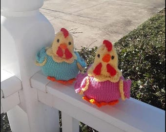 Puede incluir: Dos figuras de gallina tejidas. Una es azul, amarilla y naranja, y la otra rosa, amarilla y naranja. Tienen crestas rojas y están posadas en una barandilla blanca.