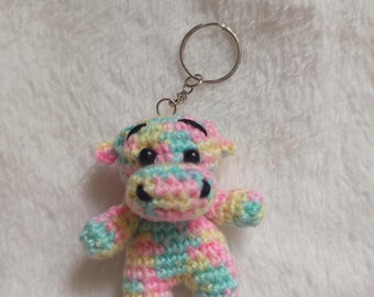 Puede incluir: Un llavero de vaca hecho a mano con ganchillo en colores pastel. La vaca es principalmente rosa, azul y amarillo, con ojos negros y una nariz negra. El llavero está unido a un anillo de metal plateado.