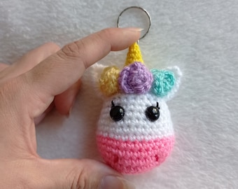 Puede incluir: Un llavero de unicornio de crochet con un cuerpo rosa, una cara blanca y un cuerno amarillo. El unicornio tiene una corona de flores con flores amarillas, moradas y azules. El llavero está unido a un anillo plateado.