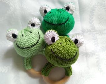 May include: Three green knitted frog toys with white eyes and black pupils attached to a wooden ring. The frogs are different shades of green.
