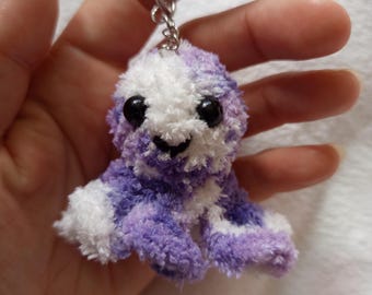 Puede incluir: Un llavero de pulpo de peluche hecho a mano en tonos morados y blancos. El pequeño y difuso pulpo tiene ojos de cuentas negras y una cara sonriente. El llavero está unido a un anillo de metal plateado.