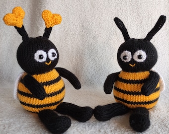 May include: Two black and yellow knitted bee toys with white wings and black eyes. The bees are sitting on a white fuzzy surface.