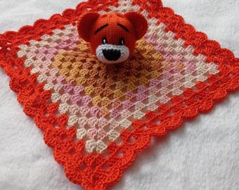 Puede incluir: Una manta de bebé de ganchillo con un oso de ganchillo naranja adjunto. La manta está hecha con hilo naranja, rosa y crema y tiene un borde festoneado.