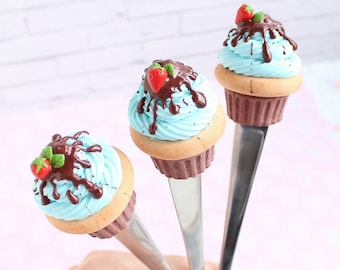 May include: Three silver spoons with blue and white frosting cupcakes topped with chocolate sauce and a red strawberry on top.