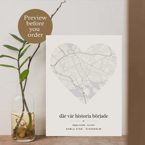 Puede incluir: Impresión enmarcada con un mapa en forma de corazón de una ciudad, con el texto "där vår historia började" y nombres y una fecha. La impresión tiene una paleta de colores neutros y se muestra con una planta en un jarrón de cristal.