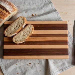 Puede incluir: Una hogaza de pan, dos rebanadas de pan, una tabla de cortar de madera con rayas alternas de madera clara y oscura y un cuchillo. El pan está rebanado y listo para servir. El cuchillo tiene un mango de madera.