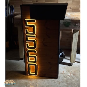 May include: A modern address sign featuring the number "5560" illuminated with a warm, yellow glow. The numbers are outlined in a bright yellow against a dark background. The sign is made of wood and has a black top.
