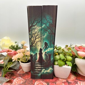 May include: A book cover with a dark forest scene. A woman in a red dress walks towards a shadowy figure on a bridge. The background is a dark green and blue with a misty effect.