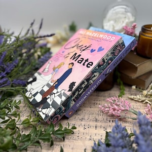 Może przedstawiać: Dwie książki w miękkiej oprawie ułożone jedna na drugiej. Górna książka jest różowa z tytułem "Check & Mate" i nazwiskiem autorki "Ali Hazelwood" w białym kolorze. Dolna książka jest niebieska z tytułem "Check & Mate" w białym kolorze. Książki leżą na kartce z nutami z różowymi i fioletowymi kwiatami.