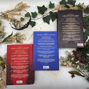 May include: Three books from the "House of Earth and Blood" and "Crescent City Series" are displayed on a background of book pages and greenery. The books have red, blue, and black covers with text. Decorative elements include a black crescent moon and floral accents.
