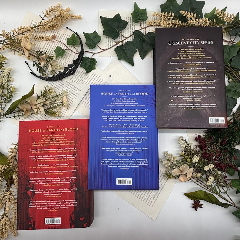 May include: Three books from the "House of Earth and Blood" and "Crescent City Series" are displayed on a background of book pages and greenery. The books have red, blue, and black covers with text. Decorative elements include a black crescent moon and floral accents.