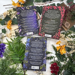 May include: Three hardcover books with floral designs and text, arranged with dried flowers and greenery. The book covers feature dark backgrounds with purple, red, and blue floral patterns. The text on the covers includes author names and book titles.