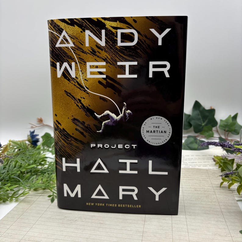 May include: A hardcover book titled "Project Hail Mary" by Andy Weir. The cover features a gold and black design with an astronaut illustration. The book is a New York Times bestseller and includes a sticker that says "The Martian".