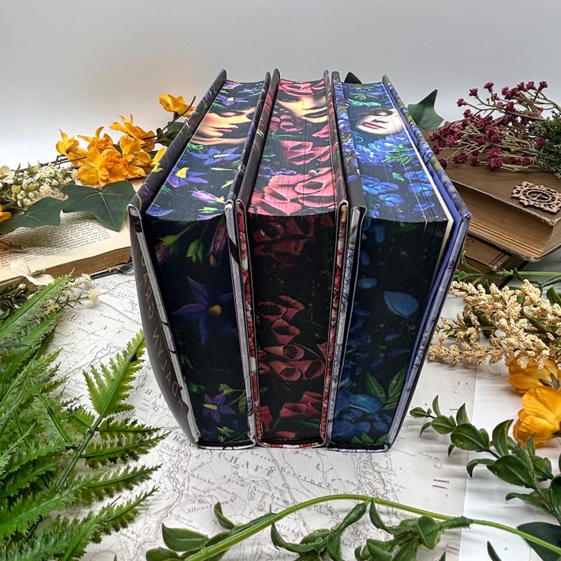 May include: Three hardcover books with floral designs on the covers. The spines are visible, showcasing a dark background with colorful flowers in shades of purple, red, and blue. The books are arranged on a map.