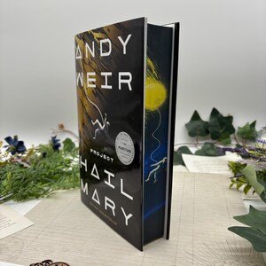 May include: A hardcover book titled "Project Hail Mary" by Andy Weir. The cover is black with white text and a graphic of an astronaut. The spine features a blue and yellow illustration. The book is a New York Times bestseller.