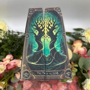 May include: A tarot card with a dark green and yellow background featuring two intertwined trees with the text "THE TWIN ALDERS" at the bottom.