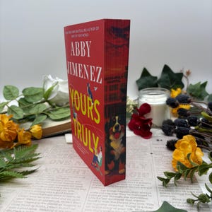 Könnte beinhalten: Ein rotes Hardcover-Buch mit dem Titel "Yours Truly" von Abby Jimenez. Das Buch hat gelbe Schrift und Illustrationen. Der Buchrücken zeigt eine Hundeillustration und die Worte "Minneapolis Hospital".