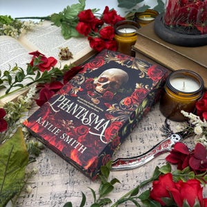 May include: A book titled "Phantasma" by Kaylie Smith with a skull and roses design on the cover. The book is surrounded by red roses, candles, and a decorative skull-shaped knife.