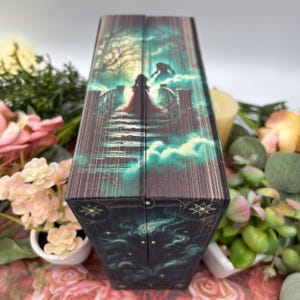 May include: A dark green and black book with a detailed illustration of a woman in a red dress walking on a bridge with a shadowy figure behind her. The book has a gold border and stars.