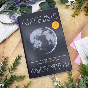 以下が含まれることがあります： アンディ・ウィアー著の「ARTEMIS」というタイトルのハードカバー本。表紙は黒で、月の銀色のイラストが描かれています。黄色のステッカーは、ニューヨーク・タイムズのベストセラーであることを示しています。本は緑と白いセーターに囲まれています。