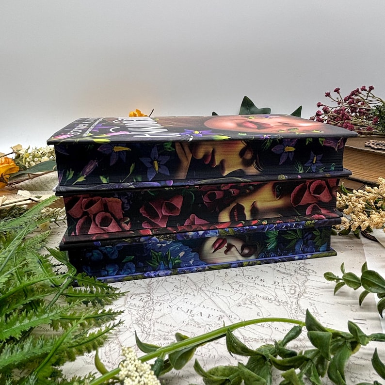 May include: Three decorative boxes stacked on top of each other. Each box features a floral design with a woman's face. The top box has the word "Witch" on it. The boxes are black with purple, red, and blue flowers.