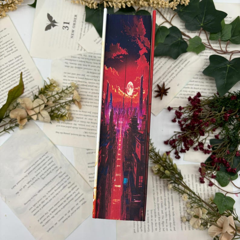 May include: A book with a vibrant cover featuring a cityscape under a red and purple sky with a full moon. The spine is visible, showcasing the artwork. The book is surrounded by pages, flowers, and greenery.