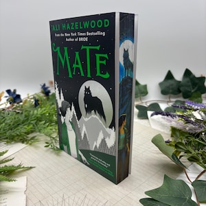 Może przedstawiać: Twarda oprawa książki zatytułowanej "Mate" autorstwa Ali Hazelwood. Okładka przedstawia czarną sylwetkę wilka na tle srebrnego księżyca, z ciemnozielonym tłem i ilustracjami postaci i drzew. Grzbiet pokazuje wilka na wzgórzu.