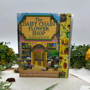 May include: A hardcover book titled "The Daisy Chain Flower Shop" by Laurie Gilmore. The cover features a colorful illustration of a flower shop with a golden retriever, flowers, and the text "The Greatest Love Is The One You Never Expected To Find."