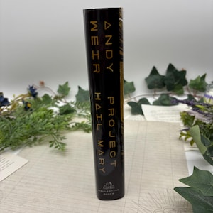 May include: A hardcover book titled "Project Hail Mary" by Andy Weir. The spine is black with gold lettering. The book is from Ballantine Books. The background includes greenery and a patterned paper.
