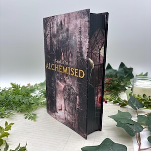 Peut inclure: Un livre à couverture rigide intitulé "ALCHEMISED" par SenLinYu. La couverture présente une illustration gothique sombre avec une église et une silhouette en manteau. La tranche du livre est noire, et le titre est en lettres dorées.