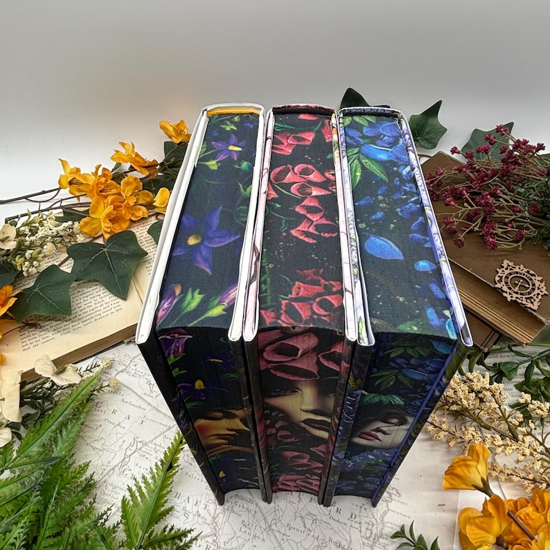 May include: Three hardcover books with floral and face designs. The book spines have silver edges and feature dark backgrounds with purple, red, and blue flowers. One book has a face design.