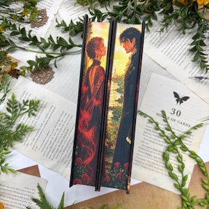 Può includere: Due libri con pittura sul taglio anteriore delle pagine. L'illustrazione raffigura una donna in abito rosso e un uomo in cappotto scuro di fronte l'uno all'altro davanti a un paesaggio al tramonto con una torre, circondati da rose rosse.