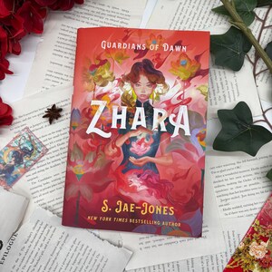May include: A book cover with a red background and the title "Guardians of Dawn" and "Zhara" in large, white letters. The cover features a woman with dark hair and a red dress holding a pink flower. The author's name, "S. Jae-Jones", is printed in white below the title.