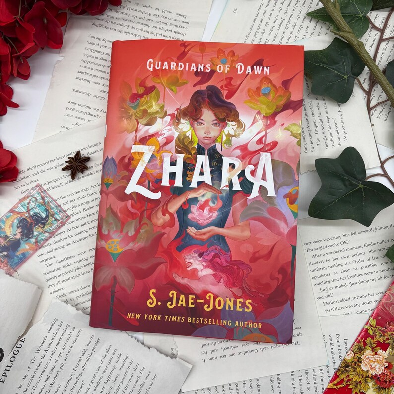 May include: A book cover with a red background and the title "Guardians of Dawn" and "Zhara" in large, white letters. The cover features a woman with dark hair and a red dress holding a pink flower. The author's name, "S. Jae-Jones", is printed in white below the title.