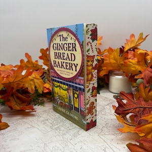 May include: A hardcover book titled "The Gingerbread Bakery" with a colorful cover featuring a bakery scene and the text "The sweetest things are worth the wait." The spine displays a cat illustration and festive decorations. Autumn leaves surround the book.