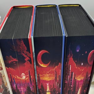 May include: Close-up of three hardcover books with black page edges. The book spines feature a futuristic cityscape design with a red and orange sky, a large moon, and a large letter "C". The book edges are red, blue, and white.