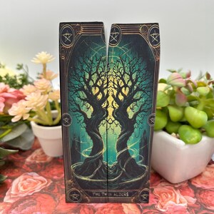 May include: A tarot card with a dark green and gold background featuring two intertwined trees with the text "The Twin Alders" at the bottom.