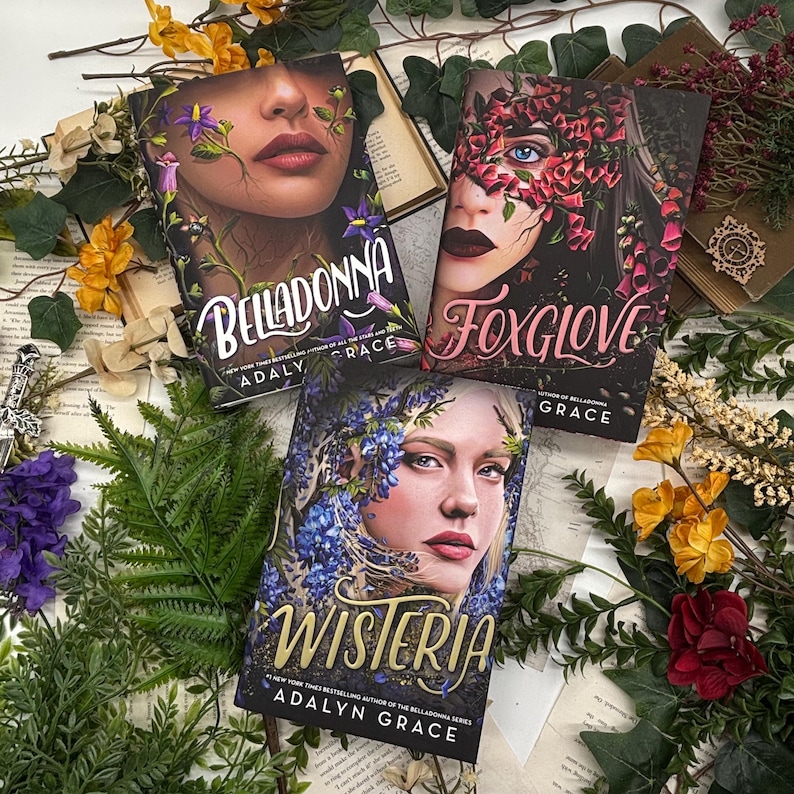May include: Three hardcover books with floral face designs. The titles are "Belladonna", "Foxglove", and "Wisteria" by Adalyn Grace. The books are surrounded by dried flowers and greenery on a vintage paper background.