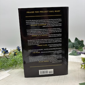May include: A hardcover book titled "Project Hail Mary" with quotes from authors and the book's ISBN. The book's cover is black with white text. The book is on a white surface with greenery in the background.