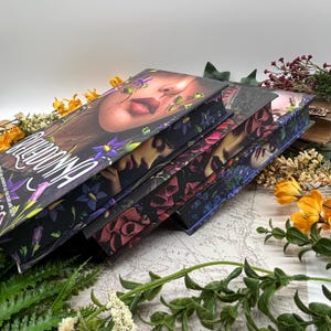 May include: A stack of hardcover books with floral and portrait cover art. The top book features a woman's face with flowers, the title "REDACTED" and author's name. The edges of the books are also decorated with floral designs. The books are surrounded by greenery and flowers.