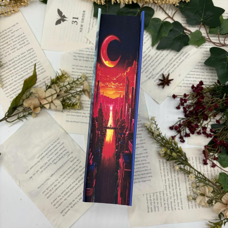 May include: A rectangular bookmark with a vibrant cityscape design. The artwork features a crescent moon in a fiery orange sky above a street lined with tall buildings. The bookmark has a blue border and is surrounded by open book pages.