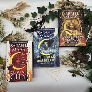 May include: Three books by Sarah J. Maas are displayed, featuring covers with illustrations and titles like "House of Sky and Breath" and "House of Flame and Shadow." The books are surrounded by decorative greenery and floral accents, creating a visually appealing arrangement.