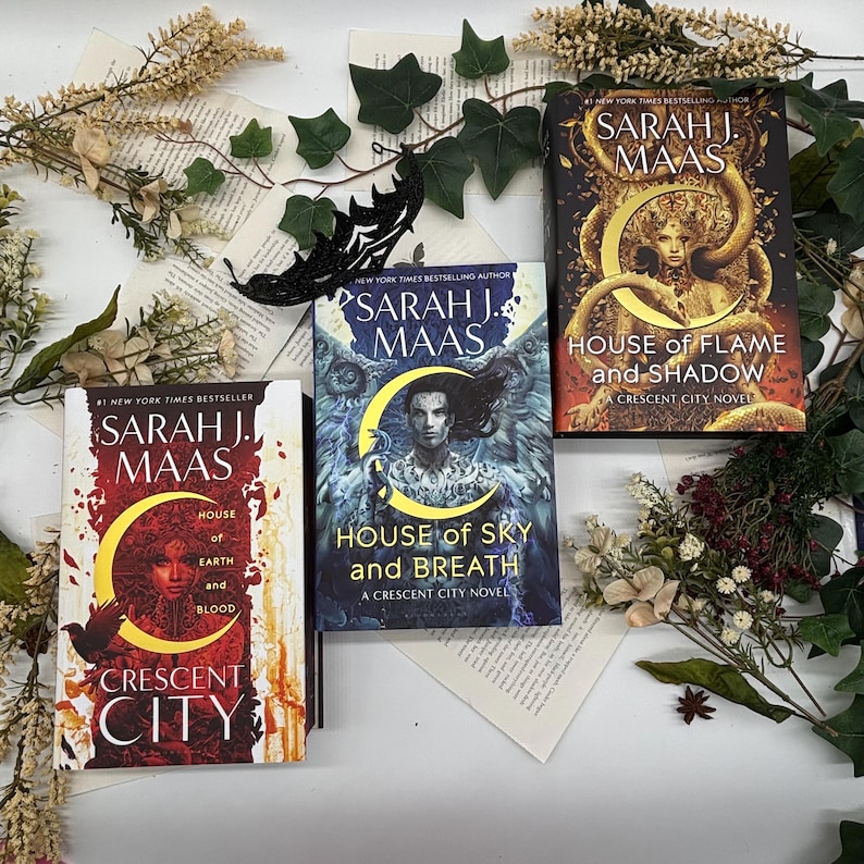 May include: Three books by Sarah J. Maas are displayed, featuring covers with illustrations and titles like "House of Sky and Breath" and "House of Flame and Shadow." The books are surrounded by decorative greenery and floral accents, creating a visually appealing arrangement.