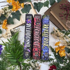 May include: Three hardcover books with floral designs and titles: "Belladonna," "Foxglove," and "Wisteria." The book spines are dark with colorful floral illustrations and the author's name, Adalyn Grace. The books are surrounded by flowers and greenery.