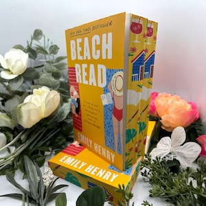 May include: A yellow hardcover book titled "Beach Read" by Emily Henry. The cover displays the title in white lettering. The spine features illustrations of beach scenes and wine glasses. The book is surrounded by flowers and greenery.
