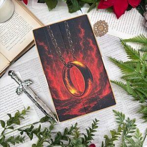 Puede incluir: Caja de madera con un anillo dorado con inscripción, suspendido por una cadena sobre un fondo rojo ardiente. La caja está rodeada por una espada decorativa, un libro abierto y vegetación, evocando un tema de fantasía.