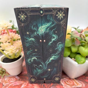 May include: A dark green and gold box with a swirling, smoky design and two glowing red eyes. The box has a gold border and stars on the front and back. The text "The Nightmare" is visible at the bottom of the box.